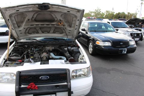 2003 Ford Crown Victoria - white Ford sedan with raised hood and push bar is parked beside a black Ford sedan in a row of show vehicles.
