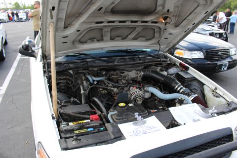 2003 Ford Crown Victoria - Underhood view of a white full-size sedan with its hood propped open at an outdoor car show, a handwritten entry card resting on the air fi…