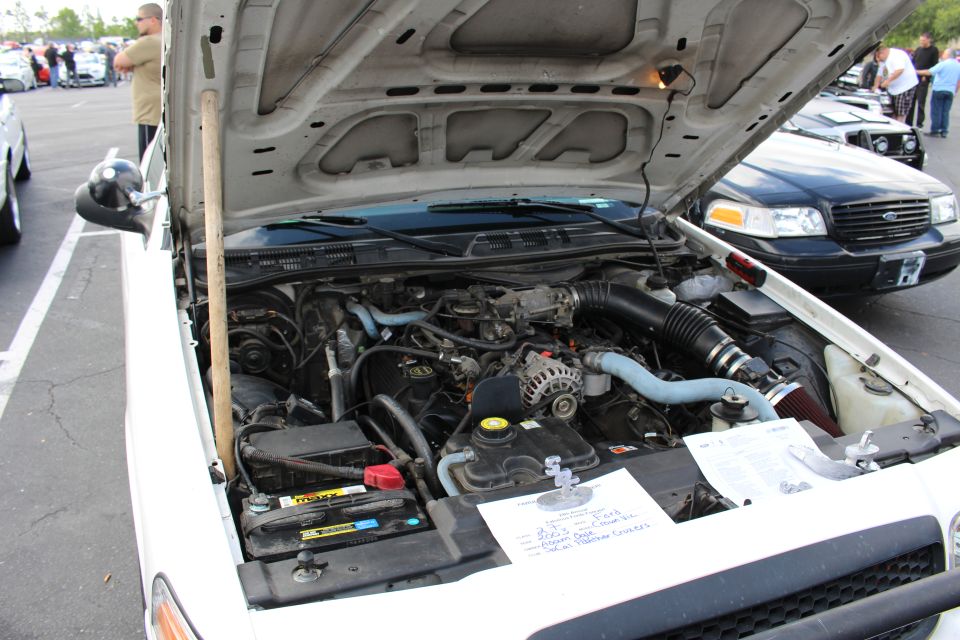 Open engine bay of a white sedan at an outdoor car show with a handwritten spec card