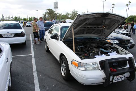 2003 Ford Crown Victoria - white Ford Crown Victoria sits with its hood open in a parking-lot car show, wearing a California plate as spectators browse nearby cars.
