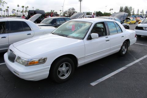 2003 Ford Crown Victoria - white Mercury Grand Marquis full-size sedan parked at an outdoor car show, framed by palm trees and rows of other show cars.