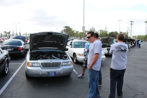 2003 Ford Crown Victoria - silver Mercury sedan displays its engine bay with the hood up and a Nevada vanity plate reading RAGE, as two men in club shirts chat beside…