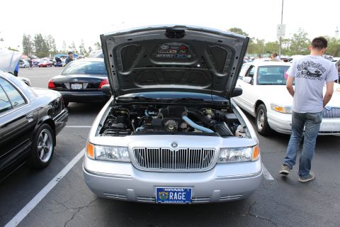 2003 Ford Crown Victoria - silver Mercury Grand Marquis sits with its hood open at a parking-lot car show, displaying a Nevada RAGE plate, a man walking past at right.