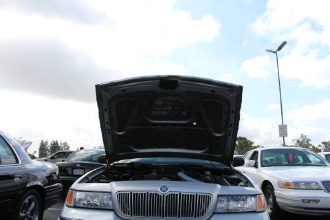 2003 Ford Crown Victoria - View under the raised hood of a silver Mercury sedan, looking up at the underhood liner and decals against a partly cloudy sky.