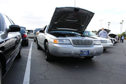2003 Ford Crown Victoria - silver Mercury sedan with its hood up and a Nevada RAGE plate sits in a row of show cars, palm trees and spectators in the background.
