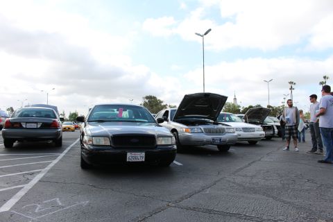 2003 Ford Crown Victoria - black Ford Crown Victoria sits at the front of a lineup of full-size sedans with raised hoods, spectators gathered at right under a grey sk…