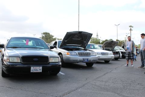 2003 Ford Crown Victoria - black Ford Crown Victoria and a silver Mercury with open hood lead a row of show sedans, with onlookers standing to the right.