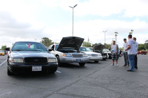 2003 Ford Crown Victoria - black Crown Victoria and several open-hood full-size sedans line a parking lot as a group of men talk beside the display.