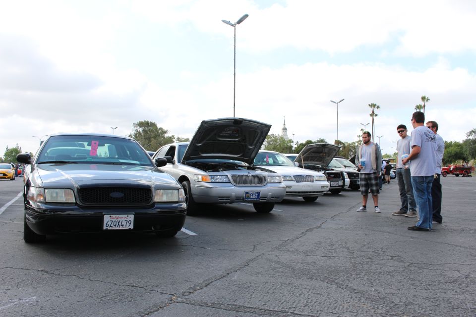 Row of Panther-platform sedans with open hoods, spectators chatting at right