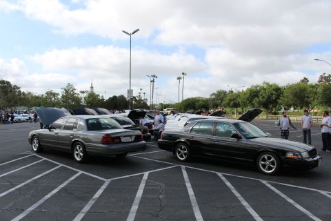 2003 Ford Crown Victoria - grey full-size sedan and a black sedan sit angled in marked parking spaces at a car show, with open-hood cars and palm trees behind.