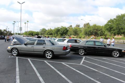 2003 Ford Crown Victoria - grey full-size sedan with its hood up sits beside a black sedan in a parking-lot car show, rows of cars and trees in the background.