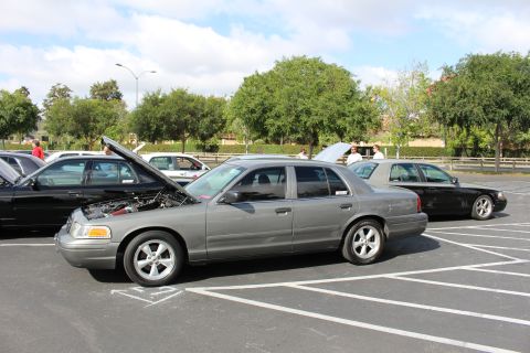 2003 Ford Crown Victoria - gray four-door full-size sedan sits with its hood open beside a black sedan in a tree-lined parking lot.