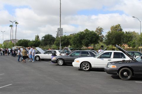 2003 Ford Crown Victoria - row of full-size sedans in black, white and gray sit with hoods open as people walk along the parking lot aisle.