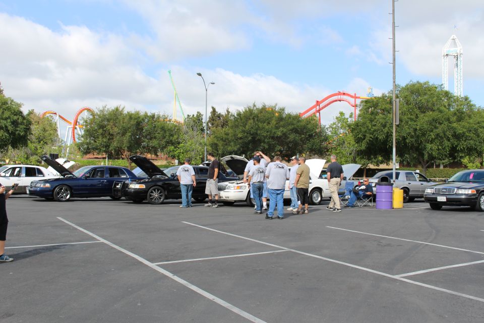 People gather around sedans in a parking lot with roller coasters behind
