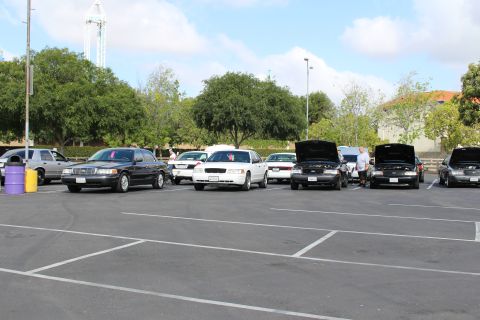 2003 Ford Crown Victoria - Black and white full-size sedans line up with hoods open across a wide empty parking lot, a drop tower in the distance.