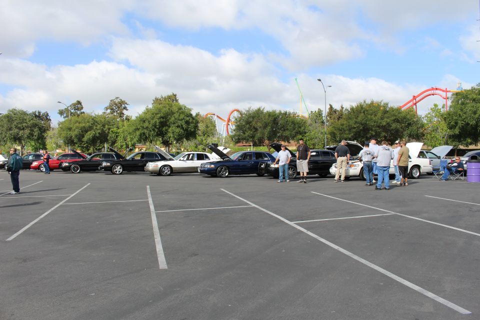 Wide view of a car meet with sedans and people and roller coasters behind
