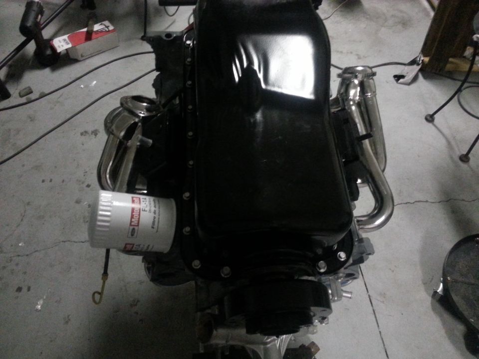 Ford V8 engine block with oil filter and chrome exhaust headers. It's sitting on a concrete floor