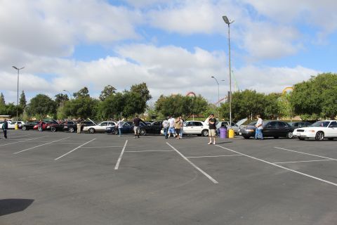 2003 Ford Crown Victoria - wide mostly empty parking lot with a distant row of sedans and scattered people, amusement park rides behind the trees.