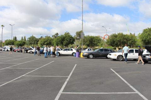 2003 Ford Crown Victoria - row of full-size sedans and onlookers fill a parking lot with a distant red roller coaster visible against the cloudy sky.