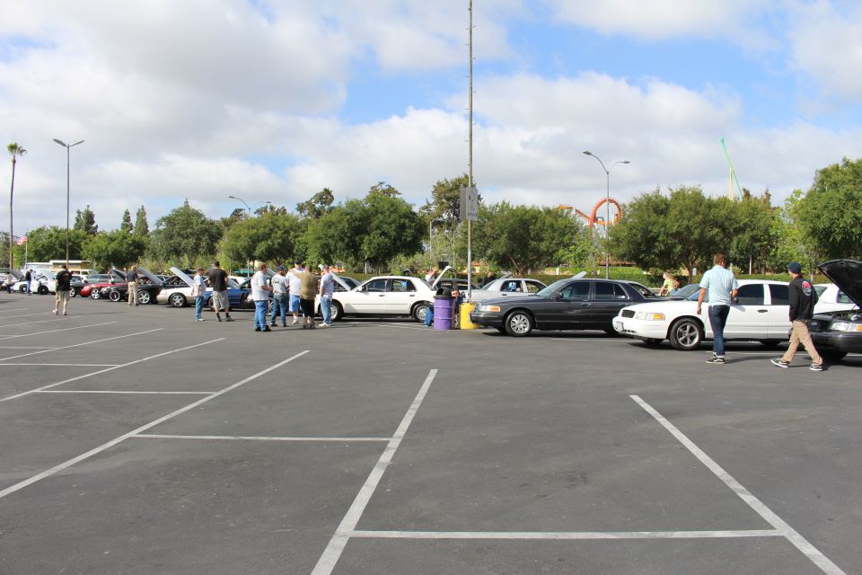 People and sedans parked in a paved lot under a cloudy sky near an amusement park ride