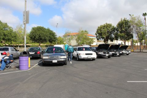 2003 Ford Crown Victoria - diagonal lineup of dark and white sedans sits with hoods open, a purple drum and a seated spectator at left.