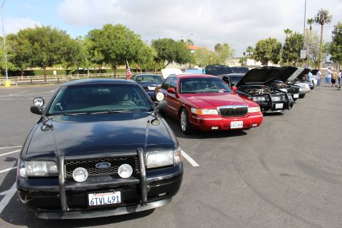 2003 Ford Crown Victoria - black Ford sedan fitted with a push bar leads a curving row of cars including a red sedan and an American flag.