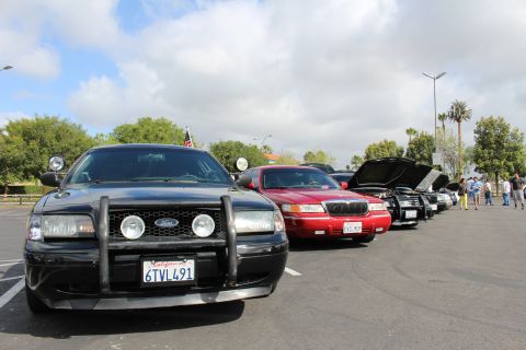 2003 Ford Crown Victoria - chrome-headlamp front of a black Ford sedan with a push bar anchors a row of parked cars under a cloudy sky.