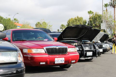 2003 Ford Crown Victoria - red sedan with a chrome waterfall grille and EVIL MGM plate sits beside dark cars displaying open hoods.