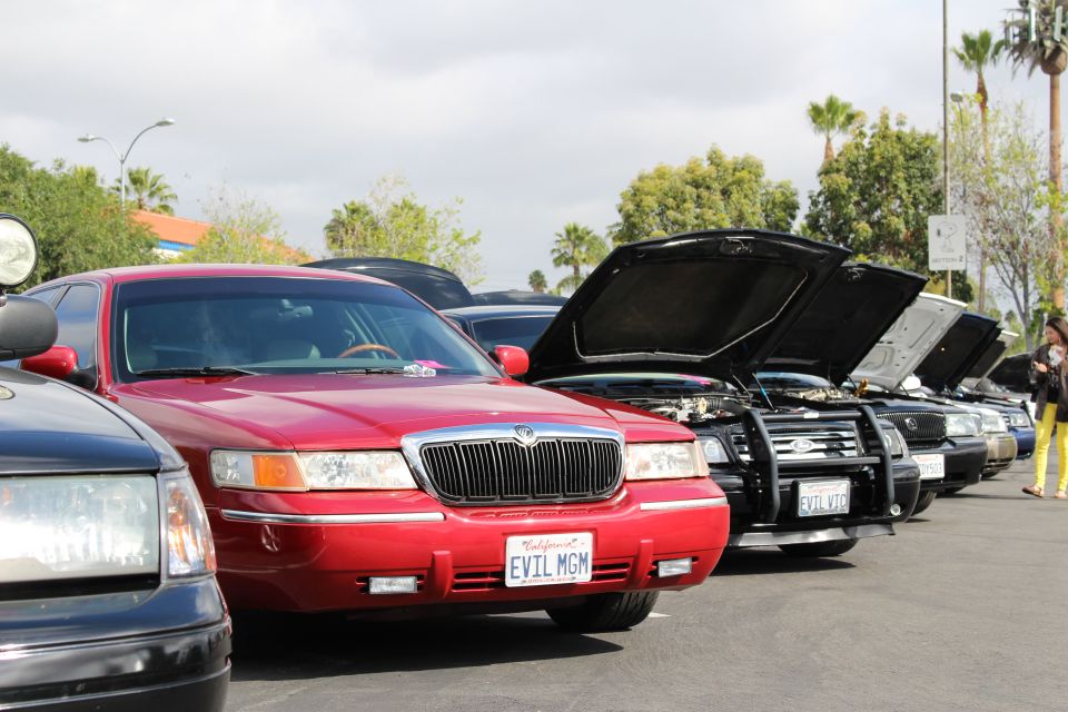 Front of red sedan with vertical-bar grille and plate EVIL MGM beside cars with open hoods