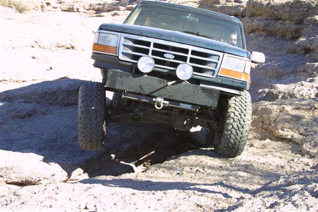 Dark green Ford truck front end crawling over rocks, head-on view with fog lights