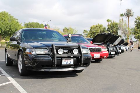 2003 Ford Crown Victoria - black Ford sedan with a brush guard and an American flag heads a row of parked cars with hoods open behind.