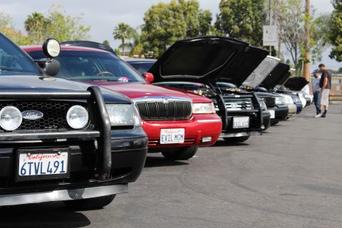 2003 Ford Crown Victoria - tight row of car front ends shows a black Ford with a push bar, a red EVIL MGM sedan, and others with hoods up.