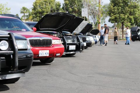 2003 Ford Crown Victoria - receding line of sedans, several with hoods open, runs past a red EVIL MGM car as spectators chat alongside.