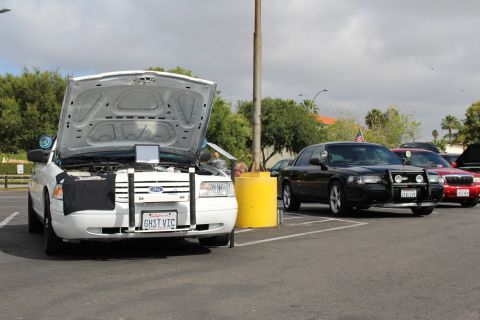 2003 Ford Crown Victoria - white Ford sedan with its trunk up and GHST VIC plate sits beside a black sedan and a yellow pole in the lot.
