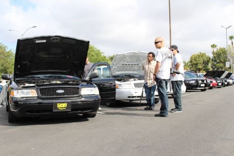 2003 Ford Crown Victoria - black Ford sedan with hood raised and a yellow plate frame sits as several men inspect the lineup of cars.