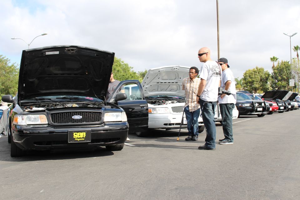 Black Ford sedan with open hood and yellow plate frame, men looking on in a lot