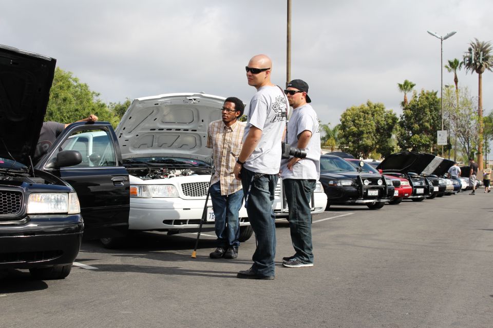 Three men standing beside sedans with open hoods in a parking-lot car meet