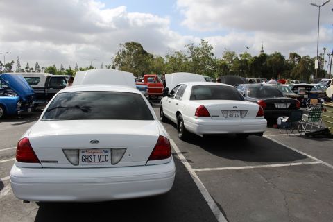 2003 Ford Crown Victoria - rear ends of two white Ford sedans, the near one plated GHST VIC, sit among many cars in a packed lot.