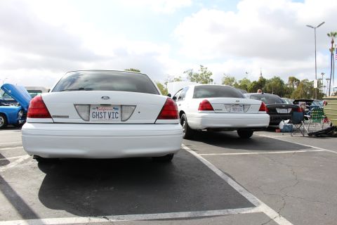 2003 Ford Crown Victoria - white Ford sedan plated GHST VIC is shown from behind next to a second white Ford in the parking-lot meet.