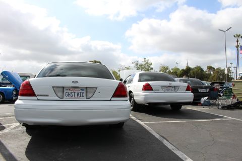 2003 Ford Crown Victoria - white Ford sedan parked at an outdoor car show, its rear California plate reading GHST VIC, with another white sedan beside it.