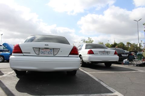 2003 Ford Crown Victoria - rear of a white Ford sedan displaying a GHST VIC California license plate, parked among other cars under a cloudy sky.