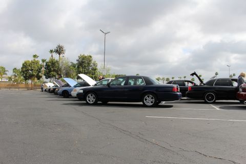 2003 Ford Crown Victoria - dark blue sedan parked in a lot among a row of vehicles with hoods open at a car gathering under an overcast sky.