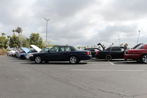 2003 Ford Crown Victoria - dark blue sedan sits in a row of cars with open hoods and trunks at an outdoor show on an overcast day.