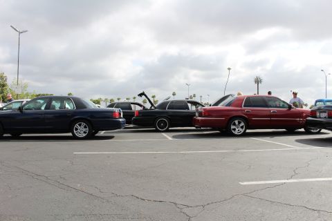 2003 Ford Crown Victoria - row of sedans including dark blue and dark red cars parked in a lot, with people standing among them under cloudy skies.