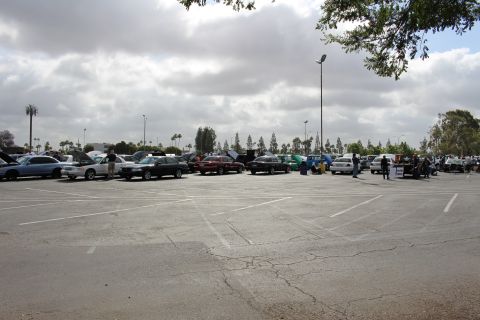 2003 Ford Crown Victoria - wide expanse of empty parking spaces leads to a distant row of show cars and a canopy under a cloudy sky.