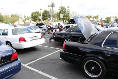 2003 Ford Crown Victoria - Black sedans, one with its hood raised, and a white sedan sit in a busy car show lot as people gather nearby.