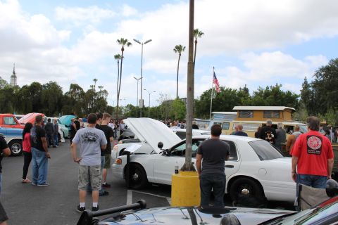 2003 Ford Crown Victoria - crowd gathers at a car show around white sedans, one with its hood up, beneath palm trees and an American flag.