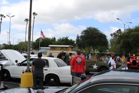 2003 Ford Crown Victoria - People stand at a car show beside a white sedan and a tan military-style vehicle, with an American flag flying behind.