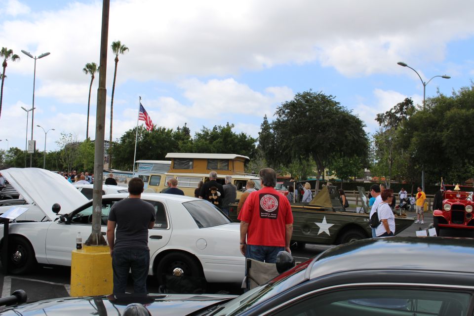 People at car show by white sedan and military-style vehicle with flag
