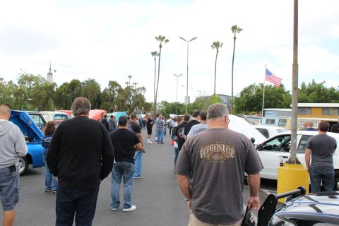 2003 Ford Crown Victoria - crowd of spectators, several in graphic tees, view show cars beneath tall palm trees and an American flag.